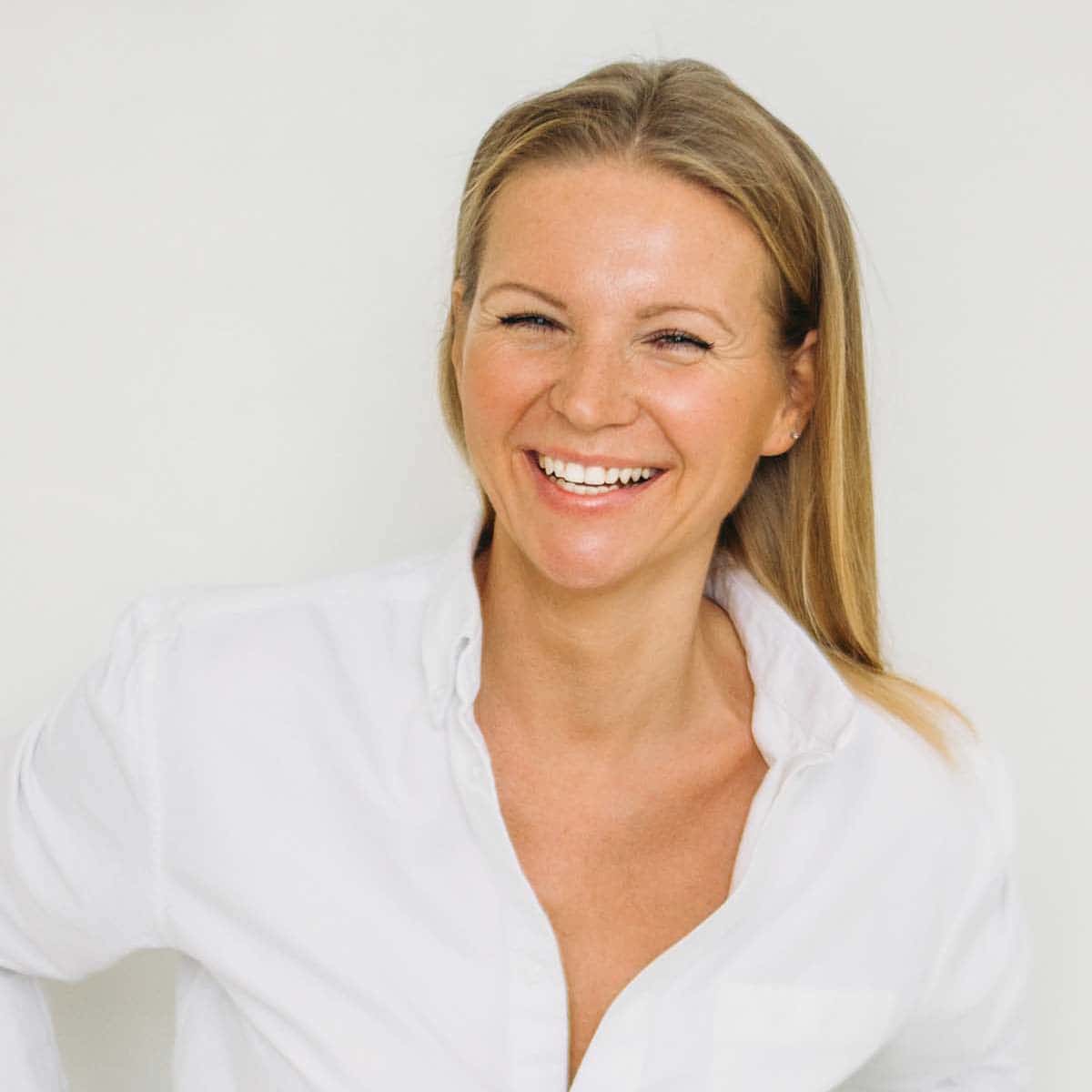 Smiling woman with straight blonde hair wearing a white button-up shirt, posing against a plain light background.