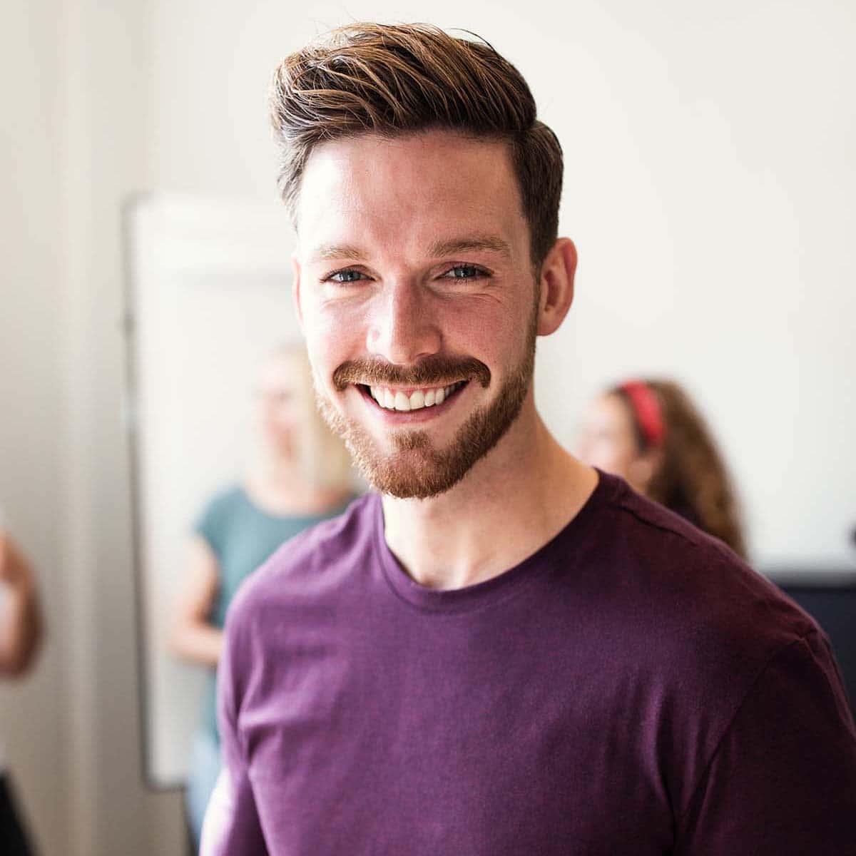 Smiling young man with styled hair and beard wearing a maroon shirt, standing indoors with people blurred in the background.