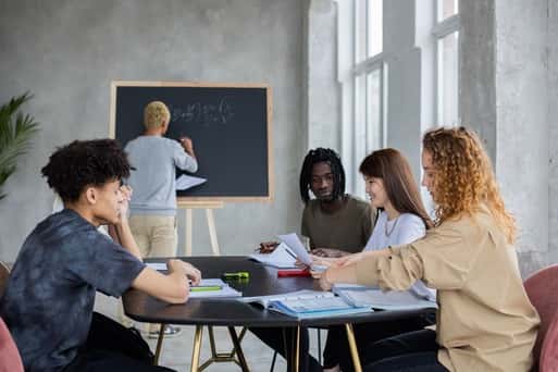 Group of students studying together at a table while one writes on a blackboard in a bright classroom.
